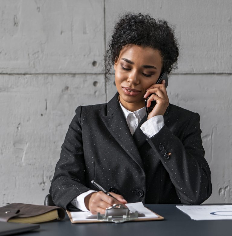 Portrait of young African American businesswoman talking on smartphone and taking notes in loft office. Concept of business communication